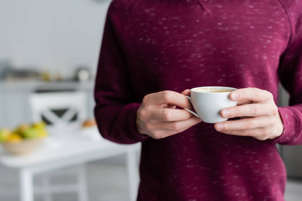 partial view of man holding cup of morning coffee in blurred kitchen - Photo, Image