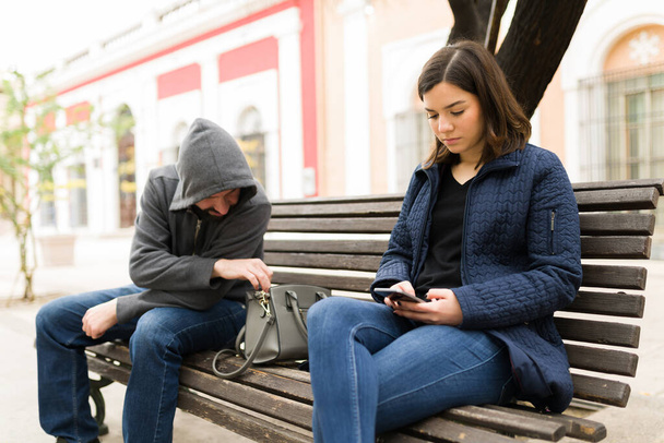 Concepto de inseguridad. Hermosa mujer joven sentada en un banco y mensajes de texto en el teléfono inteligente, mientras que un ladrón carterista su cartera  - Foto, imagen