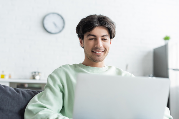 smiling brunette man working on blurred laptop at home - Photo, Image