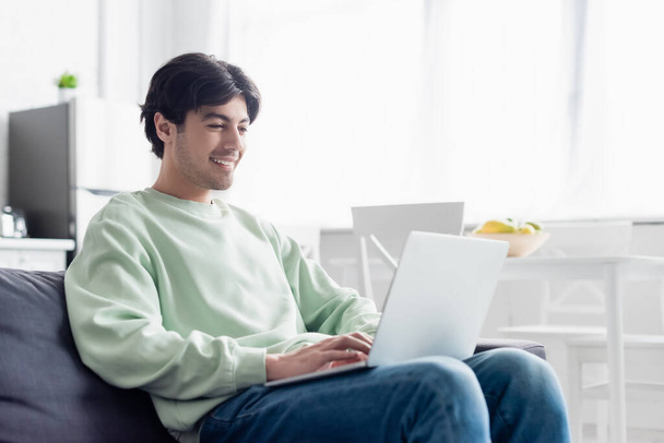 pleased man using laptop while sitting on couch at home - Фото, изображение