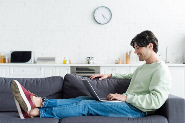 full length view of smiling man sitting on sofa with laptop at home - Photo, Image