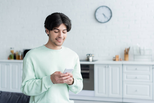 joyful man messaging on mobile phone in blurred kitchen - Photo, Image