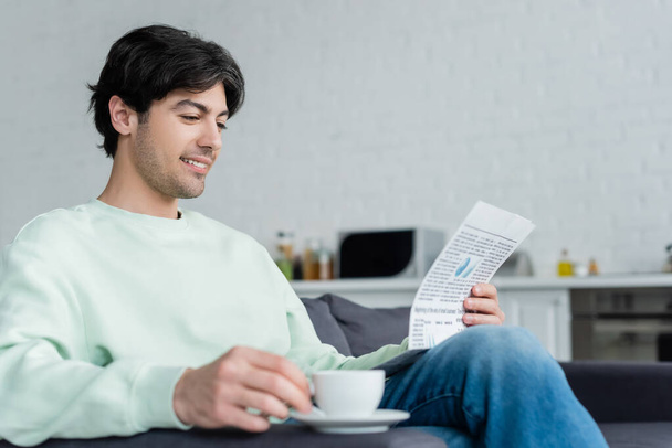 smiling man reading newspaper near blurred coffee cup on sofa - Foto, Imagen