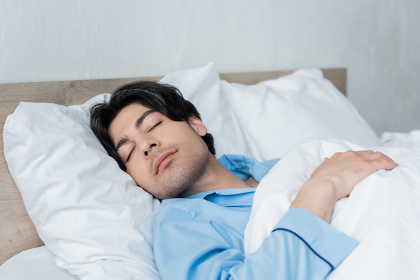 brunette man in blue pajamas sleeping on white bedding in morning - Photo, Image