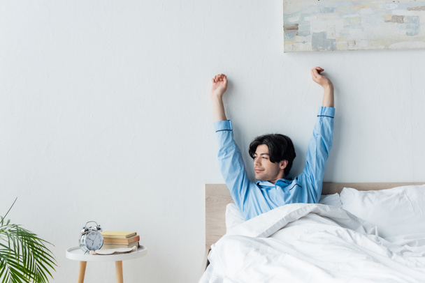 Lächelnder Mann streckt sich mit erhobenen Händen, während er im Bett neben dem Wecker sitzt - Foto, Bild