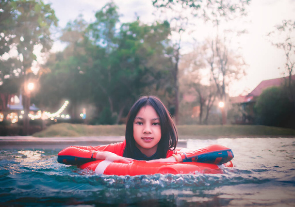 Asian pre teen girl is swimming in a floating ring in resort swimming pool in orange swimwear. - Photo, Image