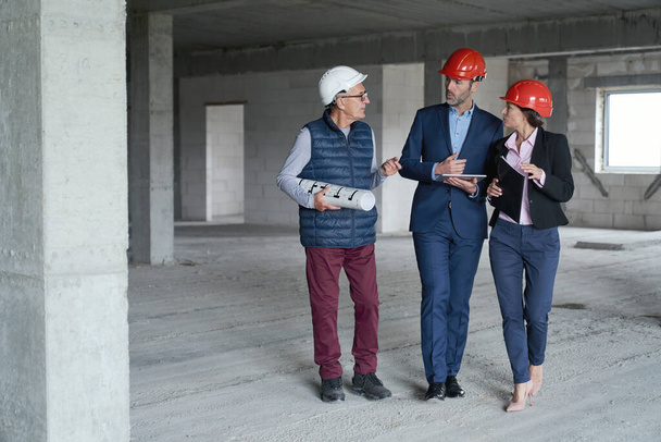 Wide image of group of caucasian engineers and investors walking and discussing on construction site  - 写真・画像