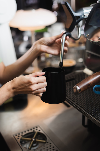 Καλλιεργημένη άποψη του barista κρατώντας κανάτα γάλακτος κοντά στον ατμό ραβδί της μηχανής καφέ στο καφέ  - Φωτογραφία, εικόνα