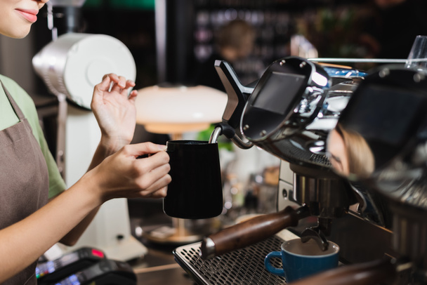 Vista recortada de un joven camarero sosteniendo jarra cerca de la varita de vapor de la máquina de café en la cafetería  - Foto, Imagen