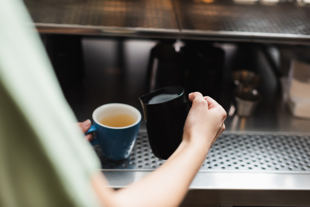 Καλλιεργημένη άποψη του barista κρατώντας κανάτα γάλακτος και κύπελλο στο καφέ  - Φωτογραφία, εικόνα