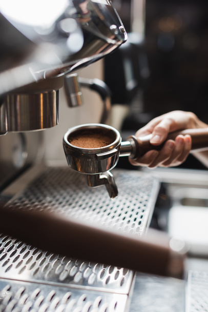 Vista recortada de portafilter de tenencia barista con café cerca de la máquina de café borrosa en primer plano en la cafetería  - Foto, imagen