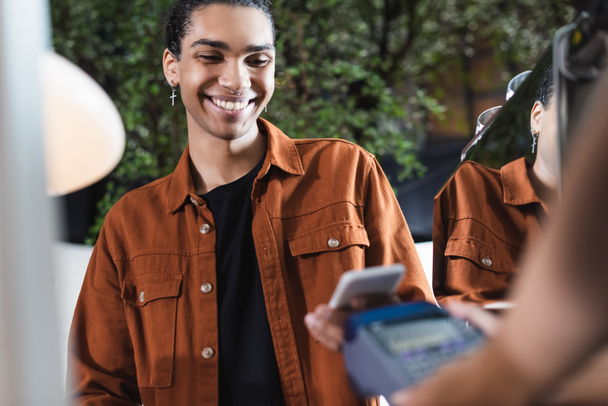 Smiling african american customer holding cellphone near blurred barista with payment terminal in cafe  - Foto, Imagen