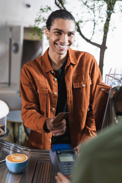 Glimlachende Afro-Amerikaanse klant houdt credit card in de buurt wazig barista met betaalterminal en koffie in cafe  - Foto, afbeelding