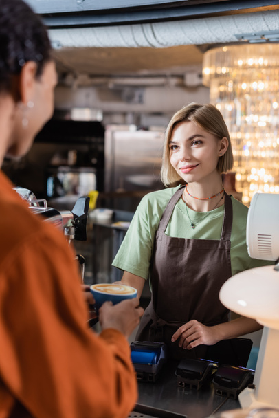 Smiling barista holding coffee near blurred african american customer in cafe  - Photo, Image