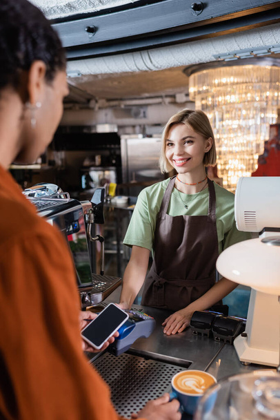 Lächelnder Barista hält Zahlungsterminal in der Nähe eines afrikanisch-amerikanischen Kunden mit Smartphone und Kaffee im Café  - Foto, Bild