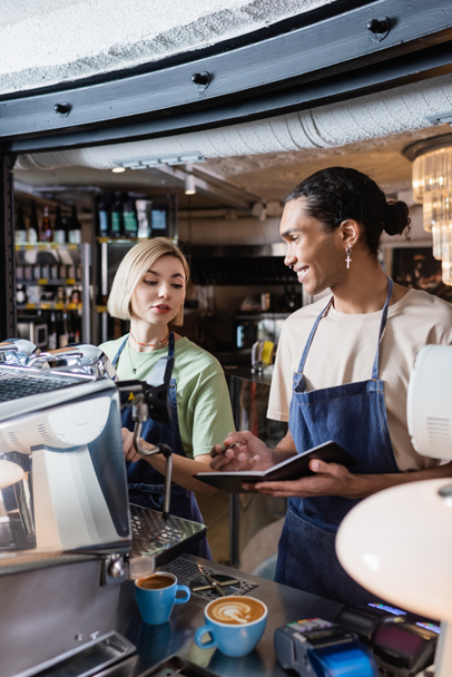 Smiling african american barista holding notebook near colleague, coffee and coffee machine in cafe  - Photo, Image