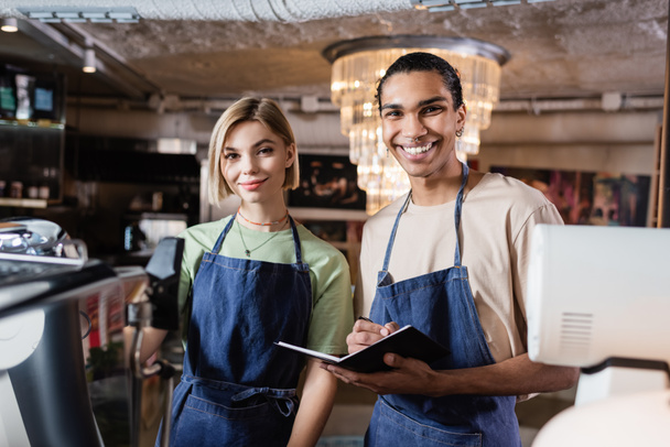 Positive multiethnic baristas with notebook looking at camera in cafe  - Photo, Image