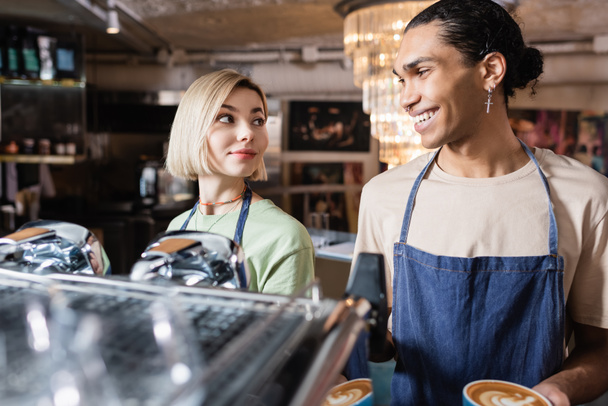 同僚とコーヒーマシンの近くにコーヒーを持っているアフリカ系アメリカ人のバリスタの笑顔カフェで  - 写真・画像