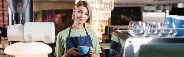 Smiling barista holding cup of coffee near coffee machine in cafe, banner  - 写真・画像