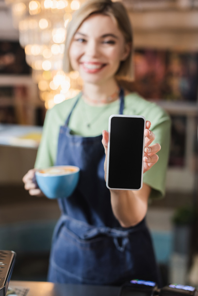 Cellphone with blank screen in hand of barista with cup in cafe  - Фото, изображение