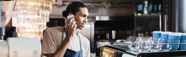 Smiling african american barista talking on smartphone near coffee machine in cafe, banner  - Foto, Imagem