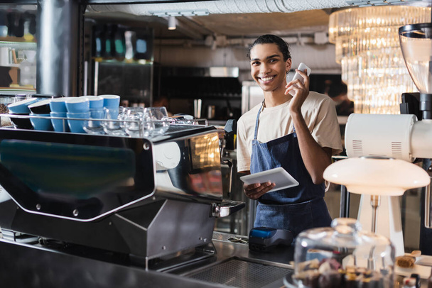 Sonriente africano americano barista dispositivos de retención cerca de la máquina de café en la cafetería  - Foto, Imagen