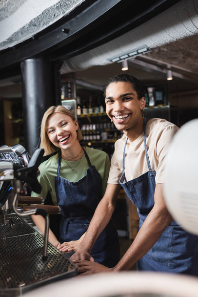 Cheerful interracial baristas looking at camera near coffee machine in cafe - Photo, Image