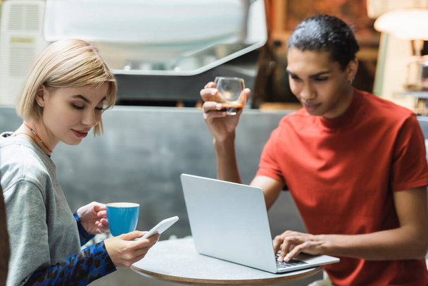 Junge Frau mit Kaffee und Smartphone in der Nähe eines afrikanisch-amerikanischen Freundes mit Laptop im Café  - Foto, Bild