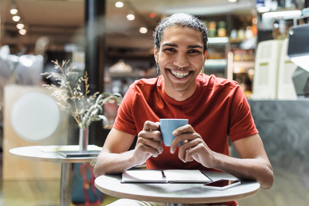Vidám afro-amerikai férfi mosolyog a kamerába, miközben kávét tart a notebook közelében a kávézóban  - Fotó, kép