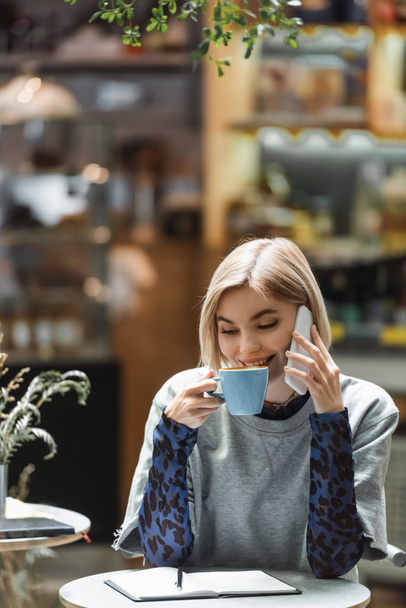 Positive woman talking on smartphone and drinking coffee near notebook in cafe  - Fotoğraf, Görsel