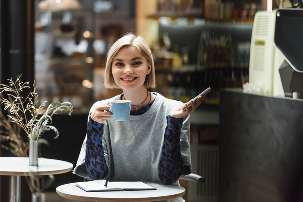 Smiling blonde woman holding cellphone and coffee near notebook in cafe  - Photo, Image