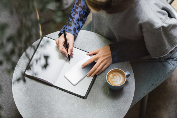 Visão geral da mulher escrevendo no notebook perto do smartphone e café no café  - Foto, Imagem