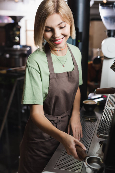Smiling blonde barista holding blurred portafilter near coffee machine in cafe  - Photo, Image