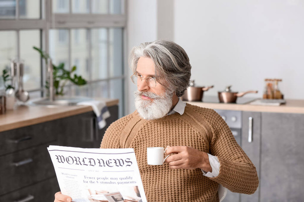 Hombre mayor con taza de café leyendo periódico en la mesa en la cocina - Foto, imagen