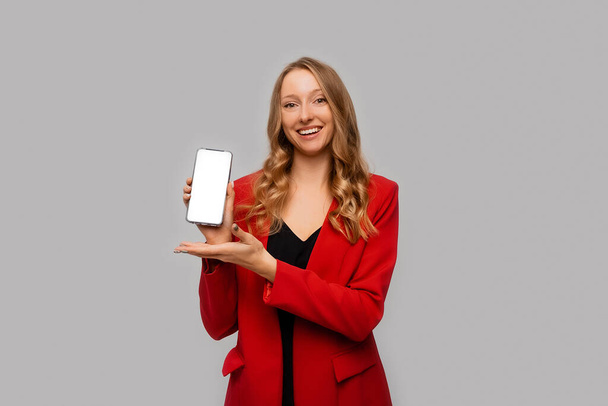 mujer de negocios hermosa y confiada mostrando la pantalla del teléfono inteligente, demostrando aplicación móvil, de pie en chaqueta roja contra el fondo gris - Foto, Imagen