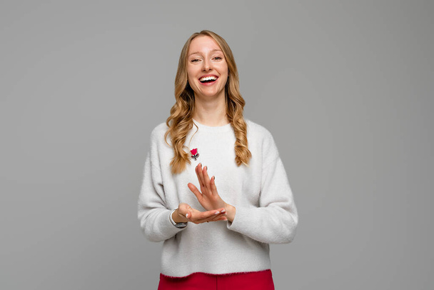 молода красива жінка відчуває себе щасливою і успішною, посміхаючись і плескаючи руками, кажучи привітання з оплесками, носить білий светр, що стоїть над сірим тлом
 - Фото, зображення