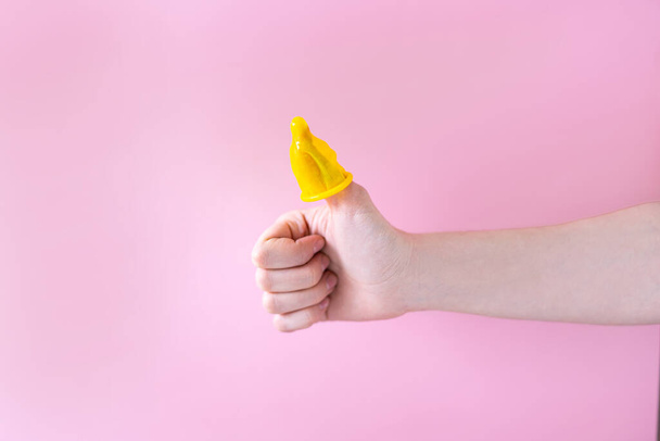 Eine Männerhand auf rosa Hintergrund hält ein Kondom. Das Kondom wird auf den Daumen gelegt. - Foto, Bild