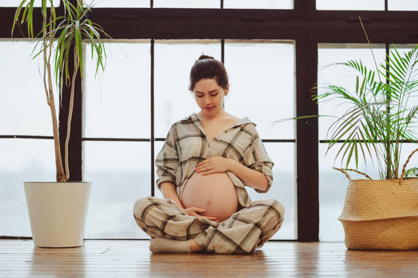 Full-length of young calm tranquil pregnant woman in pajama holding belly while sitting in lotus pose against panoramic window, expectant mother gently touching tummy. Meditation during pregnancy - Photo, Image