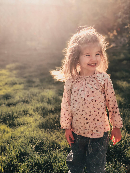 Close up photo of smiling cute little girl of 3-4 years old with blond fluffy hair stands outdoor in bright warm sunlight against backdrop of spring nature. Happy childhood concept - Foto, Imagem