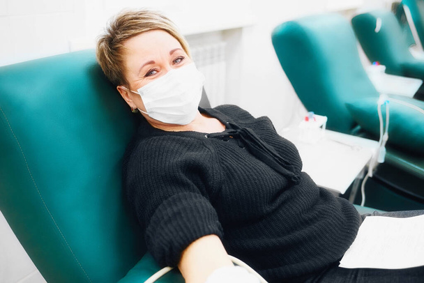 Mujer de 40-50 años con máscara médica en su cara se sienta en silla y dona sangre de la vena. Donación para enfermos - Foto, Imagen
