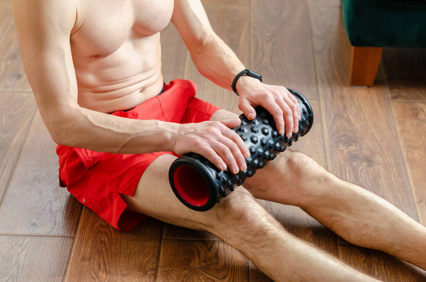 Un joven en pantalones cortos rojos está haciendo ejercicios en un rodillo de espuma, trabaja fuera de los músculos con un rodillo de masaje. Hombre atlético usando un rodillo de espuma para aliviar los músculos doloridos después de un entrenamiento. - Foto, Imagen