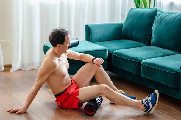 Ένας νεαρός άνδρας με κόκκινα σορτς κάνει ασκήσεις σε έναν κύλινδρο αφρού, δουλεύει από τους μυς με έναν κύλινδρο μασάζ. Αθλητικός άνθρωπος χρησιμοποιώντας ένα ρολό αφρού για την ανακούφιση των μυών πληγή μετά από μια προπόνηση. - Φωτογραφία, εικόνα