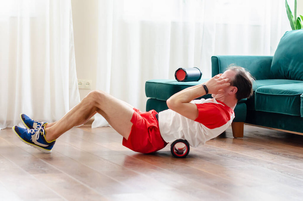 Ένας νεαρός άνδρας με κόκκινα σορτς κάνει ασκήσεις σε έναν κύλινδρο αφρού, δουλεύει από τους μυς με έναν κύλινδρο μασάζ. Αθλητικός άνθρωπος χρησιμοποιώντας ένα ρολό αφρού για την ανακούφιση των μυών πληγή μετά από μια προπόνηση. - Φωτογραφία, εικόνα