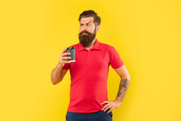 Serious bearded man in casual tshirt holding coffee cup with arm akimbo yellow background, takeaway - Foto, Bild