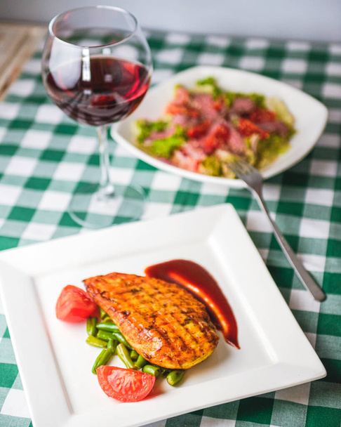 Grilled chicken breast with cherry tomatoes, green French beans, garlic, herbs, served on a white plate with a glass of red wine over wooden table. Italian cuisine concept. - Photo, image