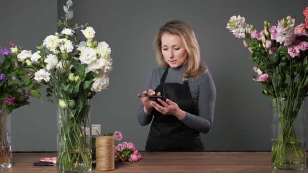 Blonde Floristin benutzt Handy in Floristik-Werkstatt - Filmmaterial, Video
