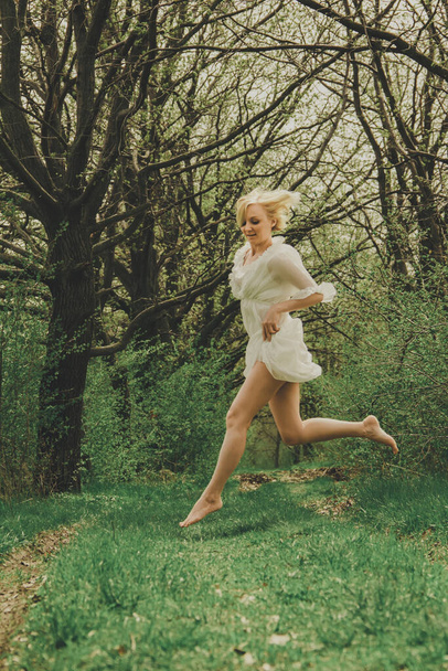 Una hermosa joven vestida de gasa blanca con un corto corte de pelo rubio corre por el camino y salta al parque. - Foto, imagen