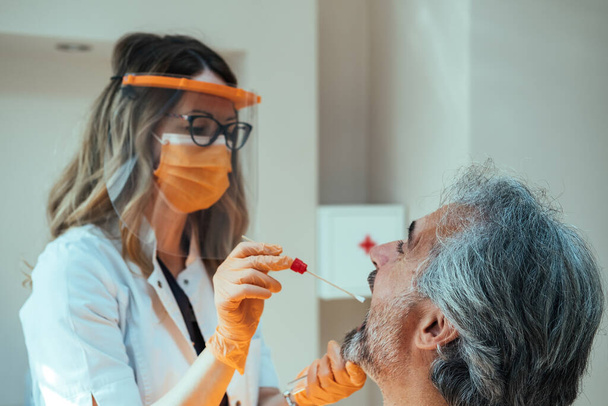 Médico femenino en ropa de trabajo protectora que toma la prueba de exudado de garganta de un paciente maduro en el hospital - Foto, Imagen