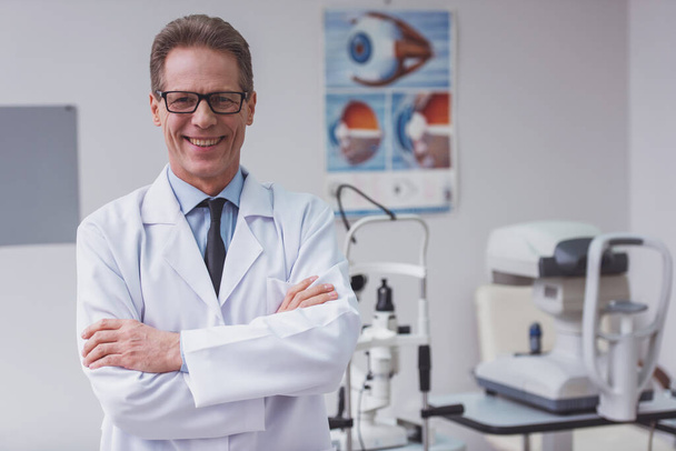 Handsome middle aged ophthalmologist looking at camera and smiling while standing in his office - Photo, Image