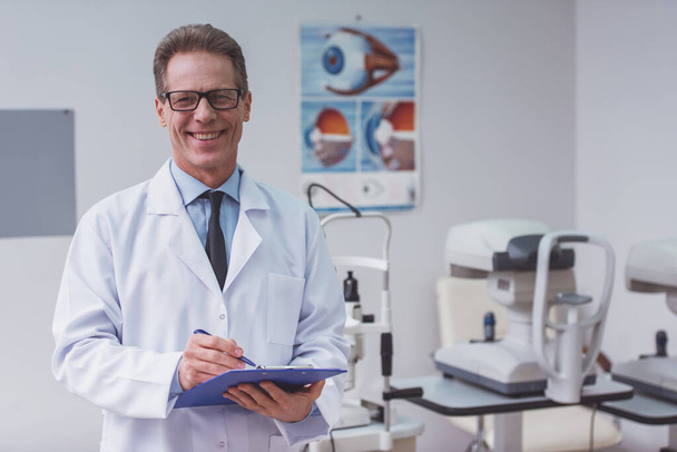 Handsome middle aged ophthalmologist making notes and smiling while standing in his office - Foto, Imagen